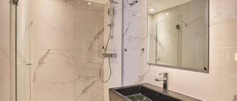 Un baño moderno con ducha de vidrio, pared con azulejos de mármol y un espejo grande sobre un lavamanos rectangular.
