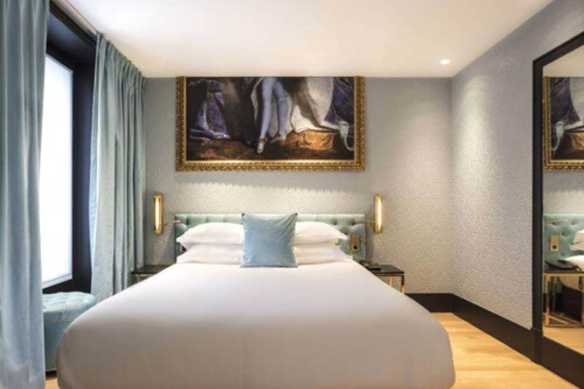 Una cama grande con edredón blanco, cabecera con diseño acolchado y dos mesitas de noche con lámparas. Hay un espejo grande en la pared y un cuadro sobre la cama.
