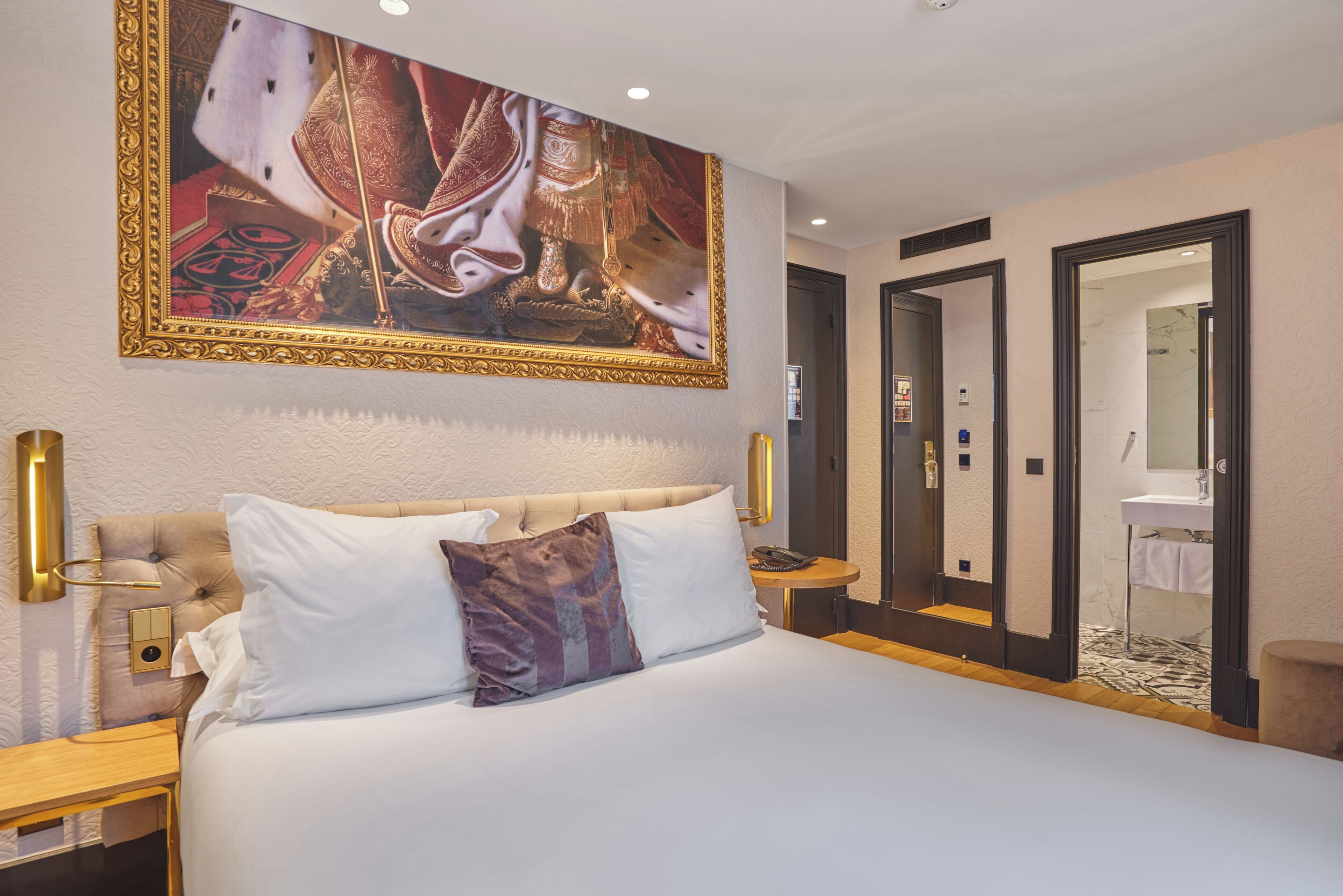 A hotel room with a large bed, a nightstand, a wall-mounted lamp, and a painting above the bed.