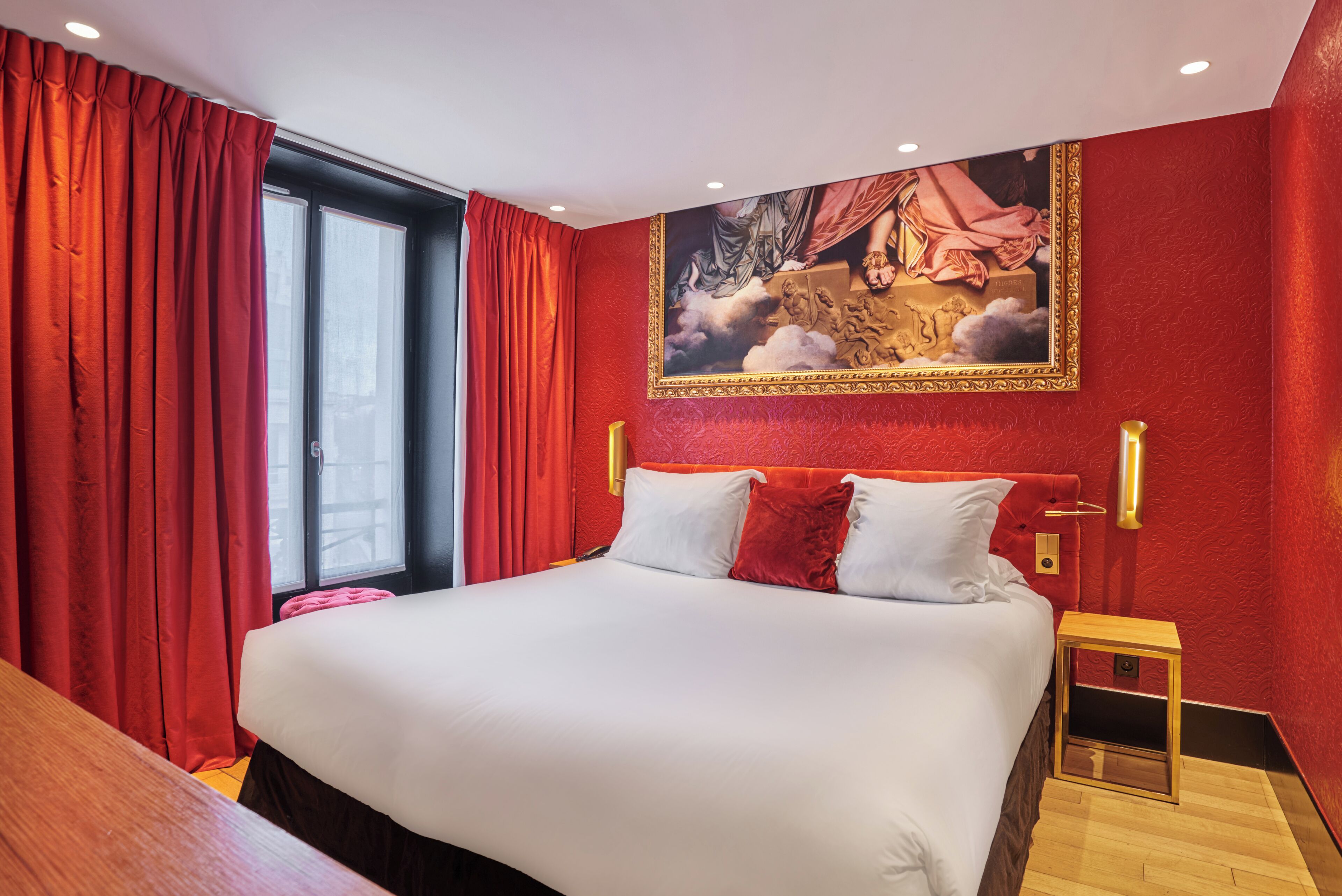 A hotel room with a large bed, red curtains, a painting on the wall, and a small wooden side table.