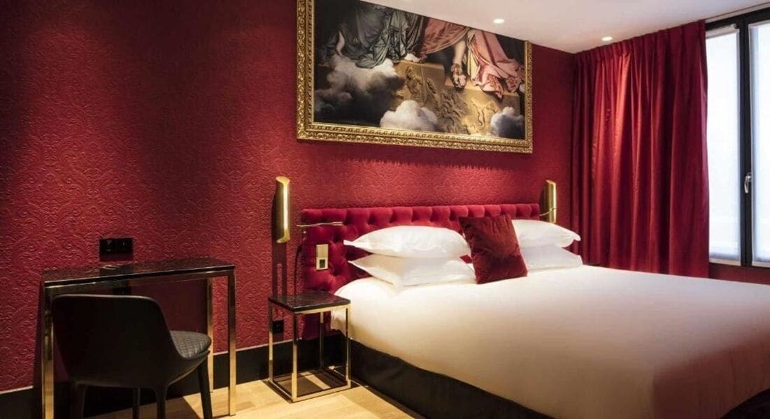 A hotel room with a large bed, a desk with a chair, a painting on the wall, and red curtains.