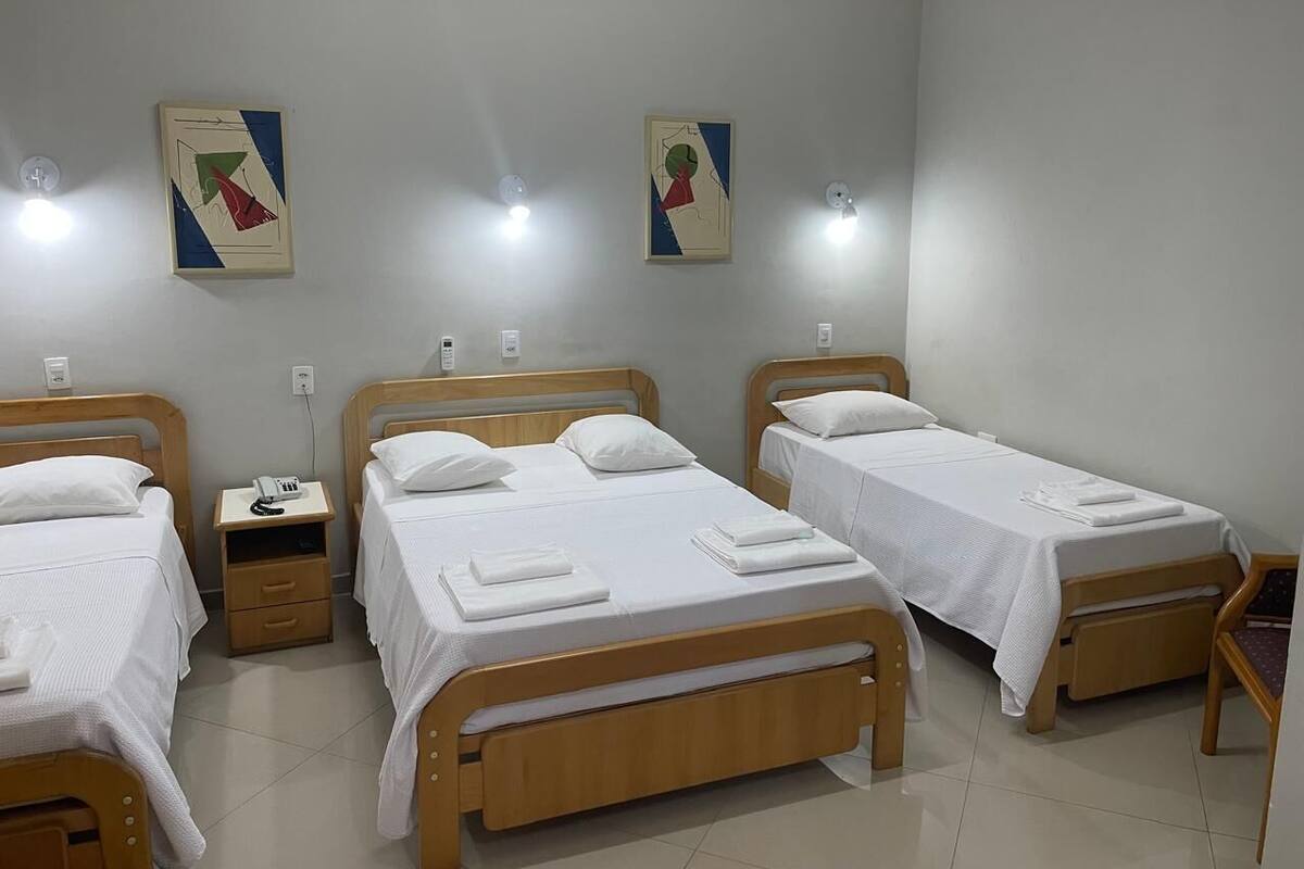 Ein Hotelzimmer mit drei Einzelbetten, jeweils mit einem Holz-Kopfteil und weißer Bettwäsche. An den Nachttischen stehen Lampen und ein Telefon. Die Wände sind schlicht gehalten, und über den Betten hängen zwei abstrakte Gemälde.