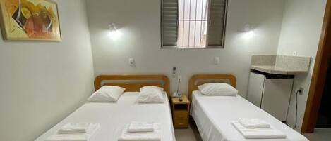 A room with two wooden beds, a small bedside table with a lamp, a window with blinds, and a painting on the wall.
