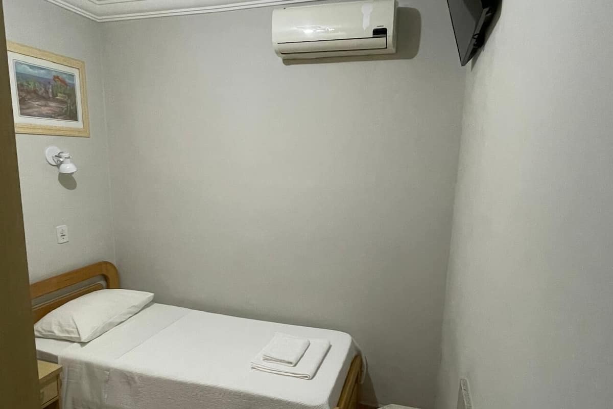 Ein kleines Zimmer mit einem Einzelbett, einem Holzkopfteil, einer weißen Bettdecke, einem auf dem Bett liegenden Handtuch, einer Wandklimaanlage und einem gerahmten Bild an der Wand.