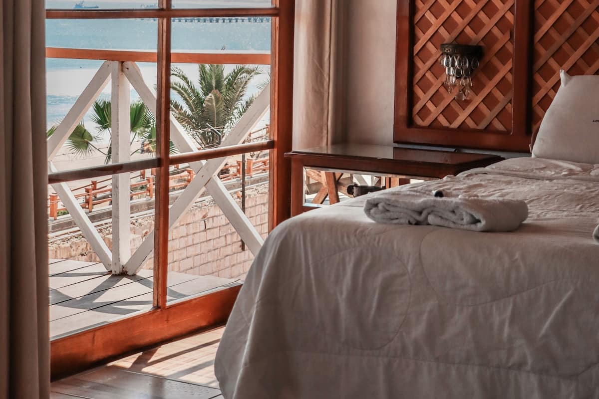 A bedroom with a large window overlooking a beach, a wooden headboard, and a neatly made bed.