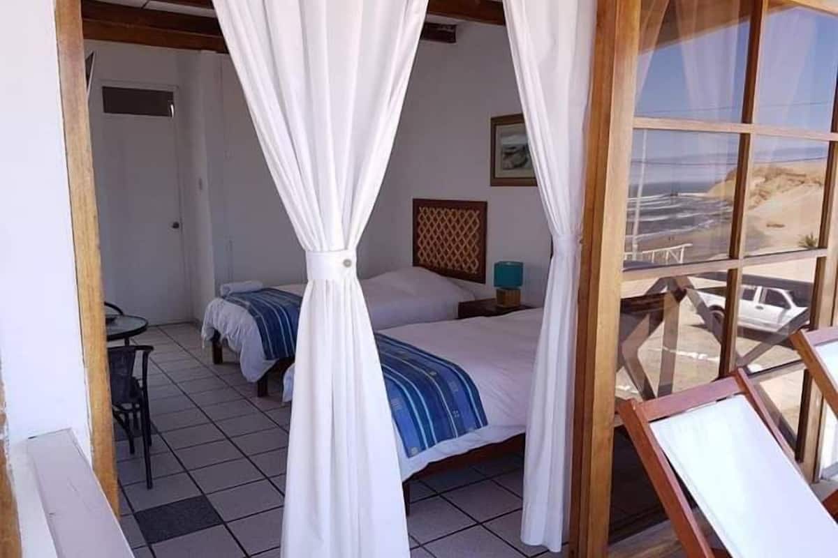 A bedroom with a bed, a nightstand, a chair, and a view of the beach through a large window.