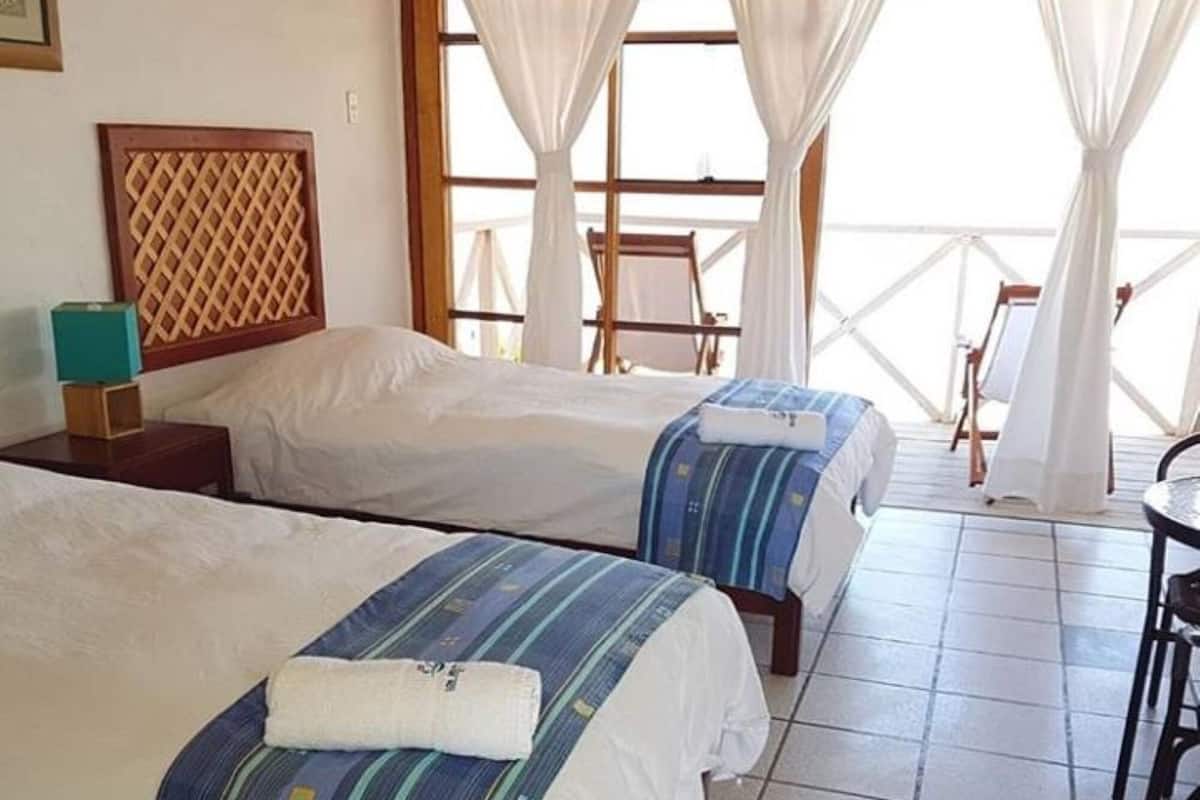 A hotel room with two beds, a wooden headboard, a small table, and a view of the outdoors through large windows.