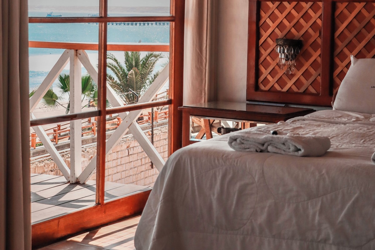 A bedroom with a large window overlooking a beach, a wooden headboard, and a neatly made bed.