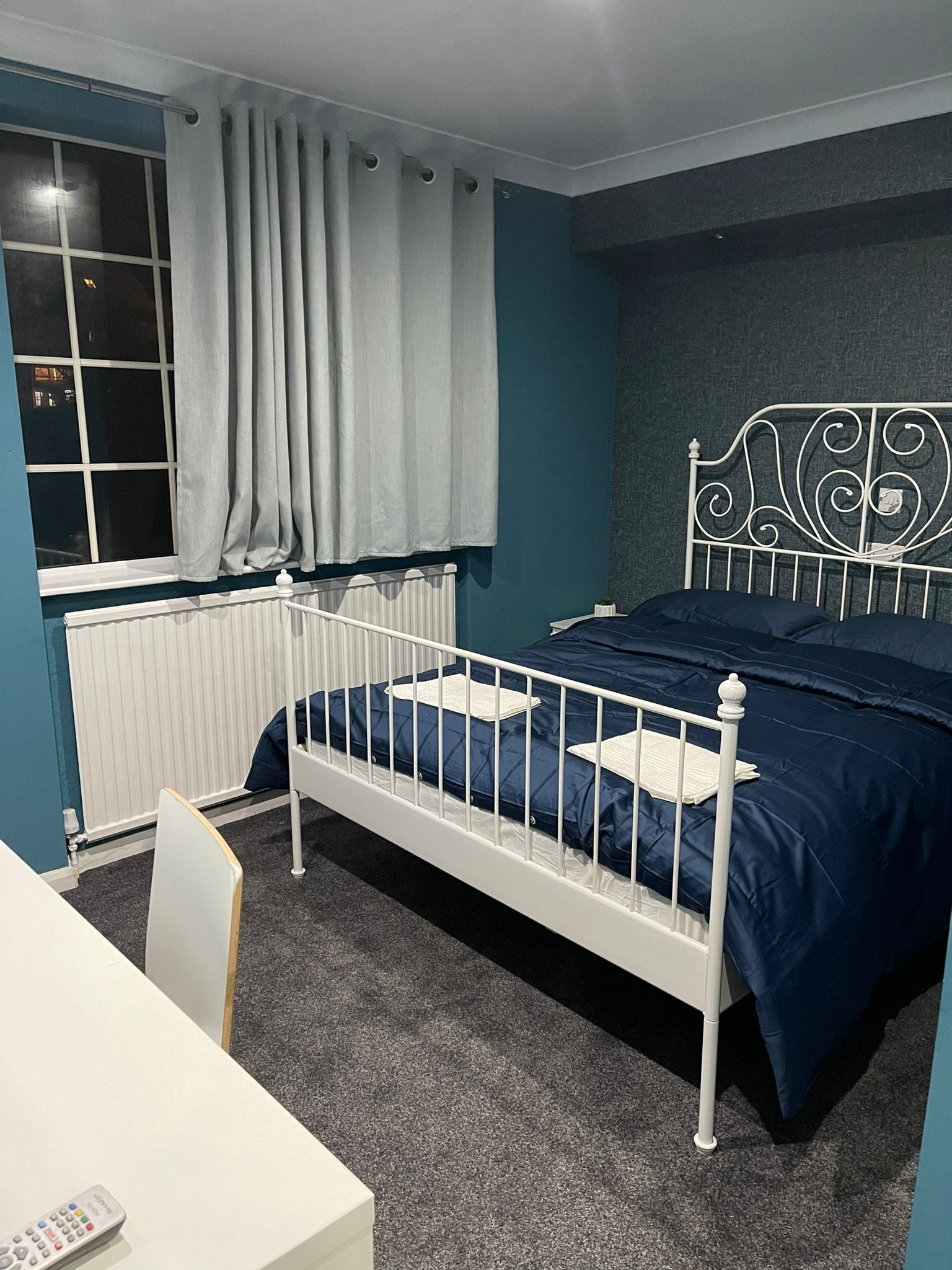 Une chambre avec un lit en métal blanc, une couvre-lit bleue, un radiateur blanc, une fenêtre avec des rideaux et un bureau blanc avec une télécommande.