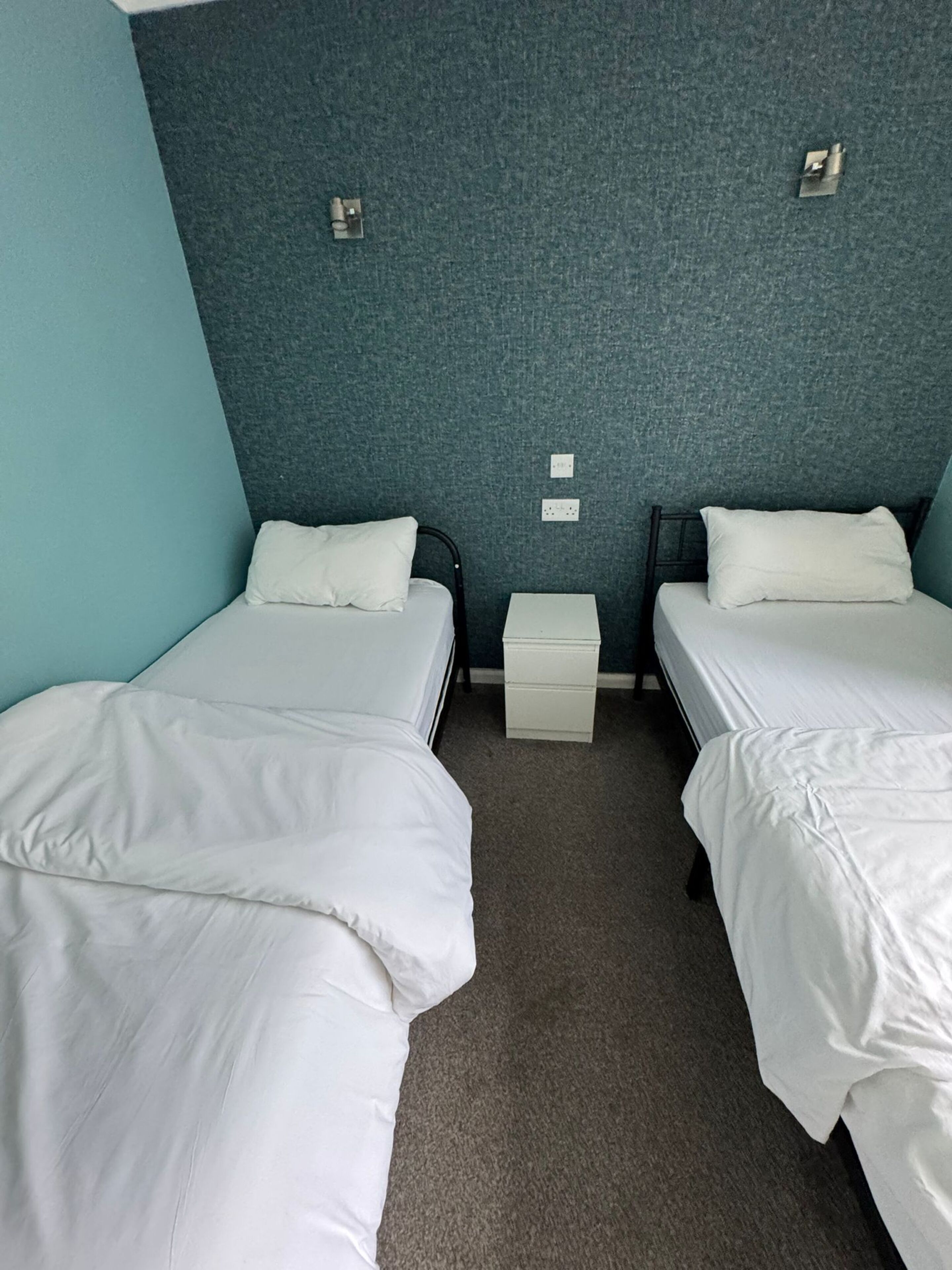 Une chambre double avec deux lits simples, une petite table de chevet et un luminaire fixé au mur.