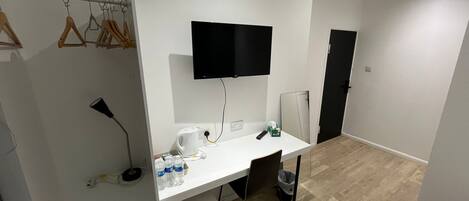 Een moderne, goed verlichte kamer met een wit bureau, een wandgemonteerde televisie en een kledingkast met houten hangers.