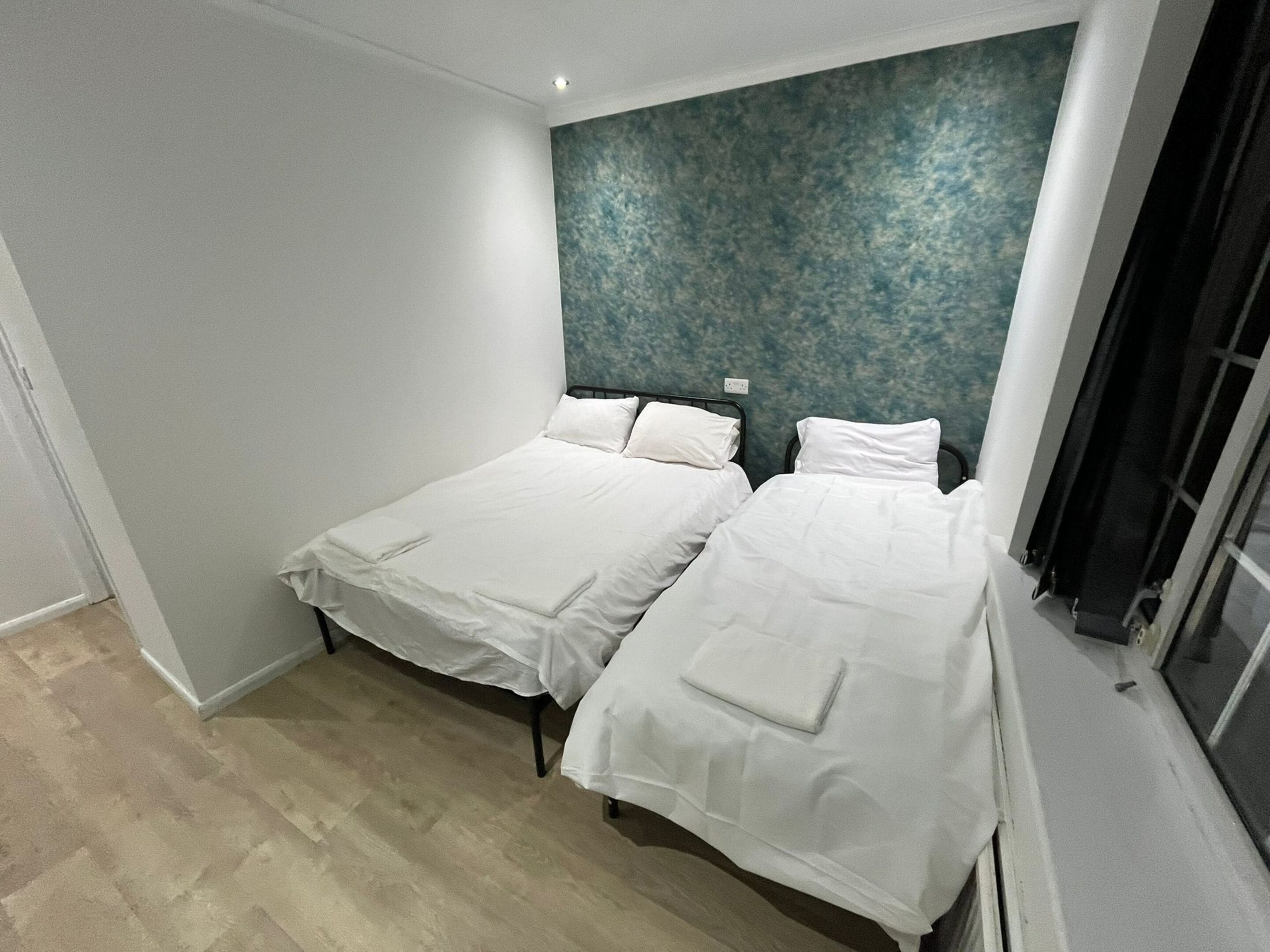 Ein Zimmer mit zwei Einzelbetten, Holzboden, einer blauen Wand mit Struktur und einem Fenster mit schwarzen Vorhängen.