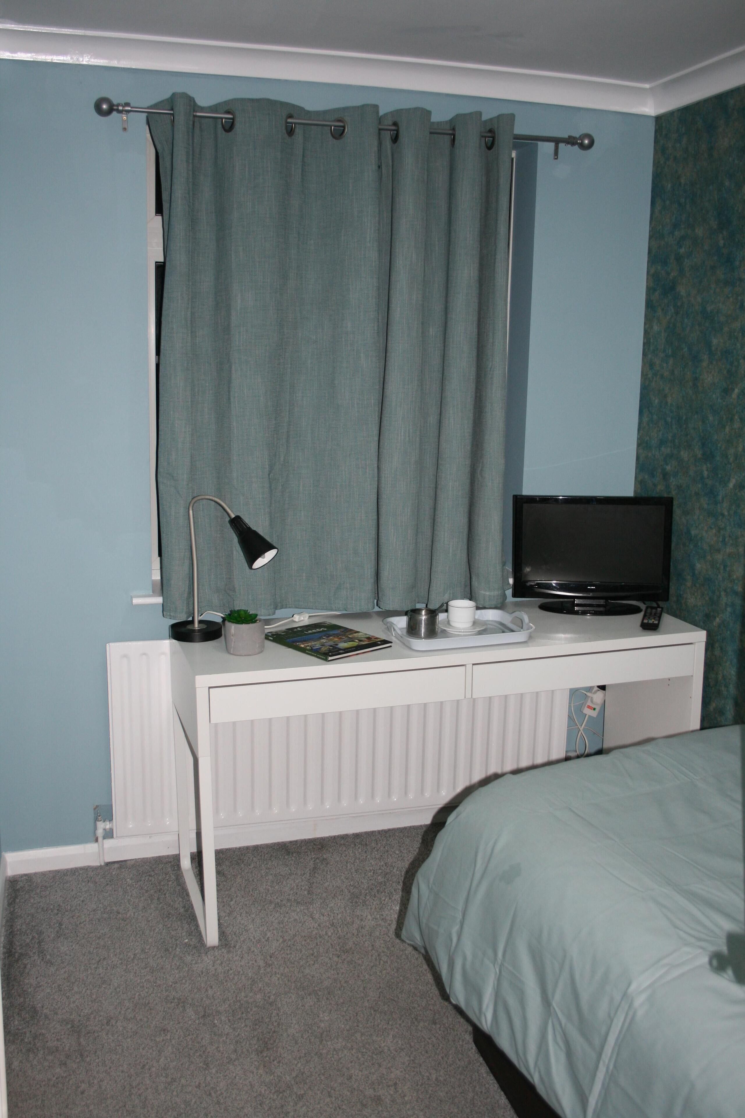 A bedroom with a bed, a desk with a lamp, a television, and a window with curtains.