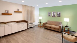 A hotel room with a sofa, a chair, a bedside table with a lamp, a wardrobe, and a window with curtains. - WoodSpring Suites Bakersfield East (Bakersfield)