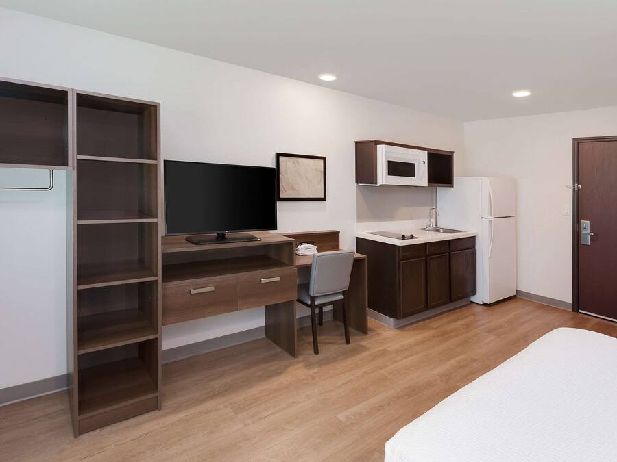 A modern, compact room with a bed, a desk with a chair, a television, a microwave, and a refrigerator.