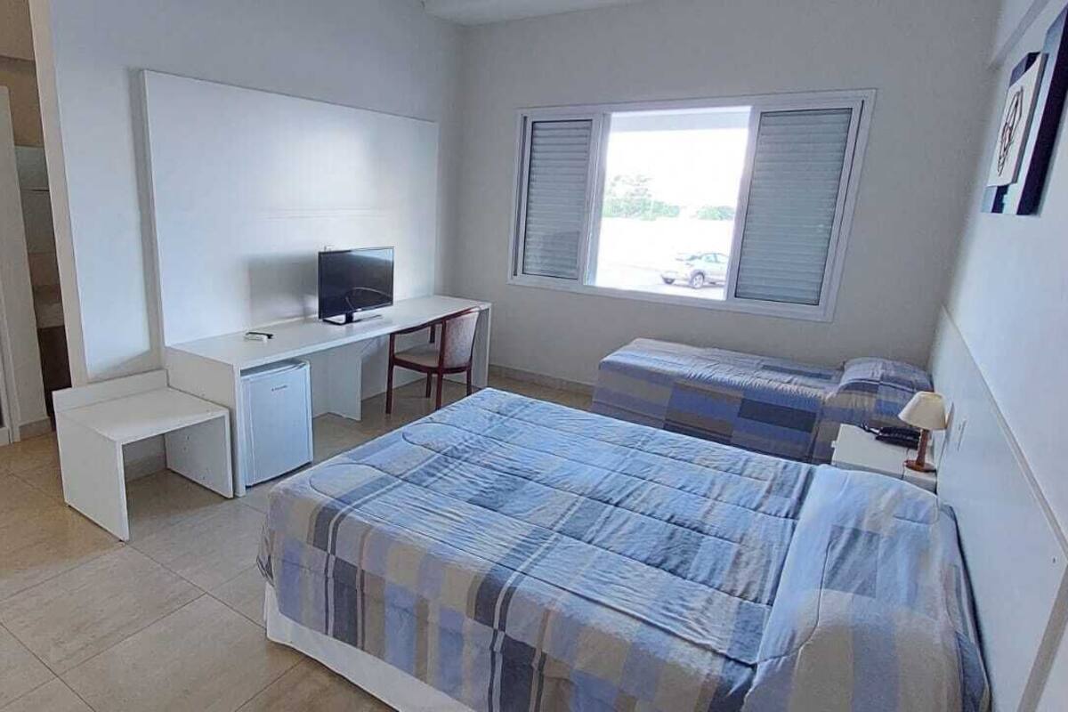 A bedroom with a bed, a desk, a chair, a TV, and a window.