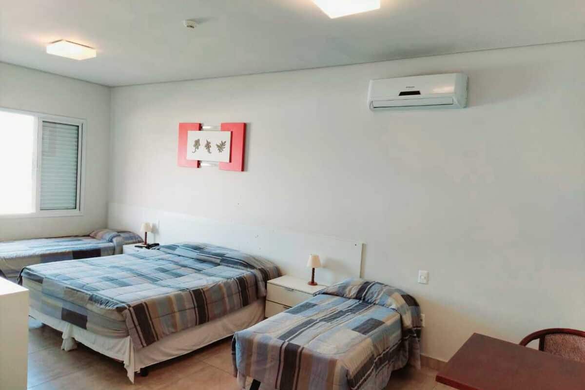 A room with two beds, a desk, and a chair. There is an air conditioning unit on the wall and a window with blinds.