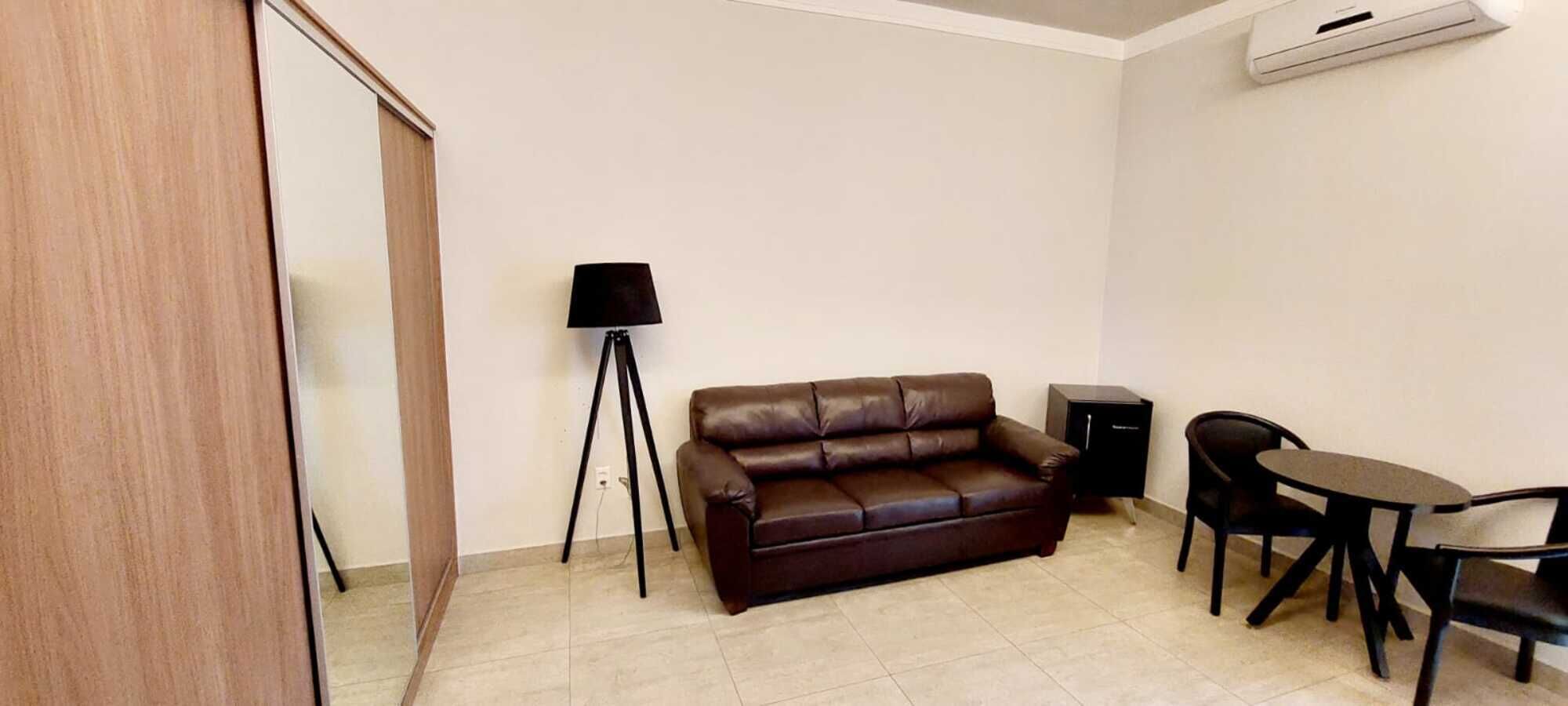 A small, furnished room with a brown leather sofa, a small round table with two chairs, a floor lamp, and a cabinet.