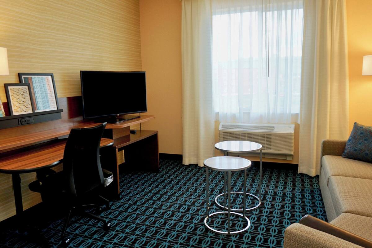Une chambre d’hôtel avec un bureau en bois, un téléviseur à écran plat, une petite table ronde, un canapé et une moquette à motifs.