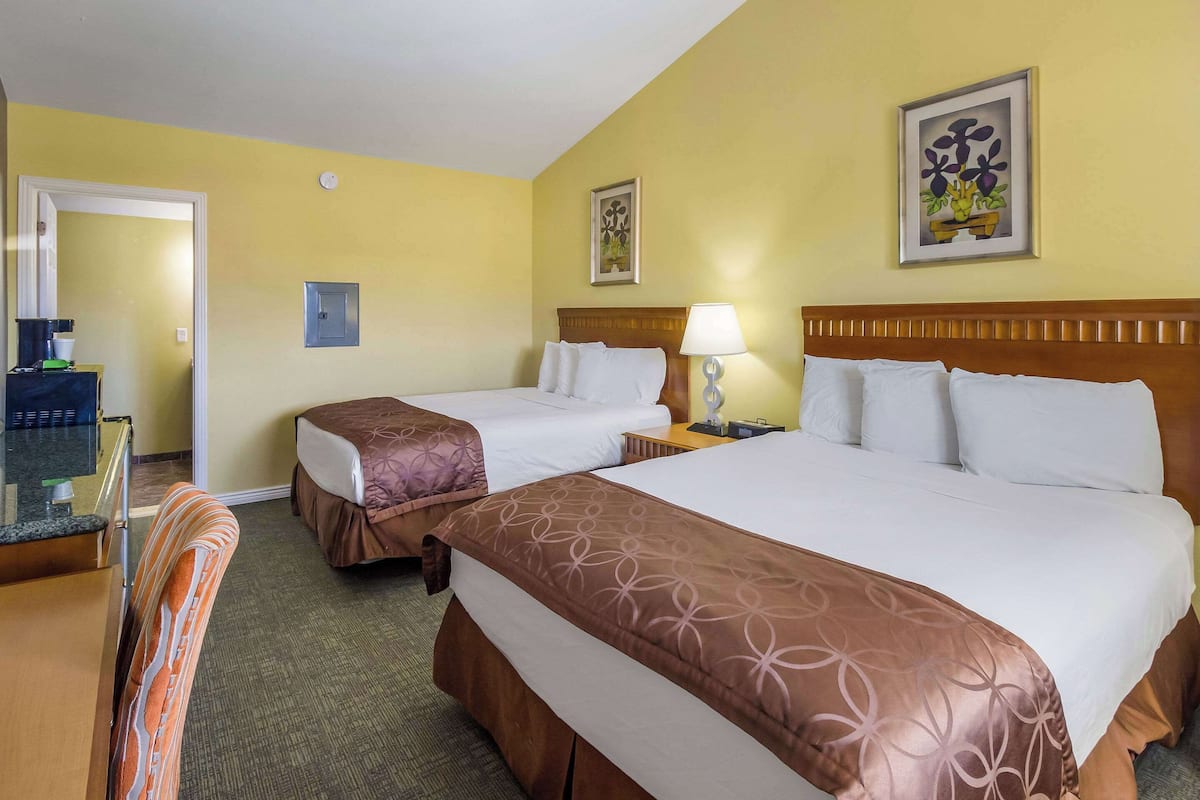 A hotel room with two beds, a desk, a chair, and a small table with a lamp.