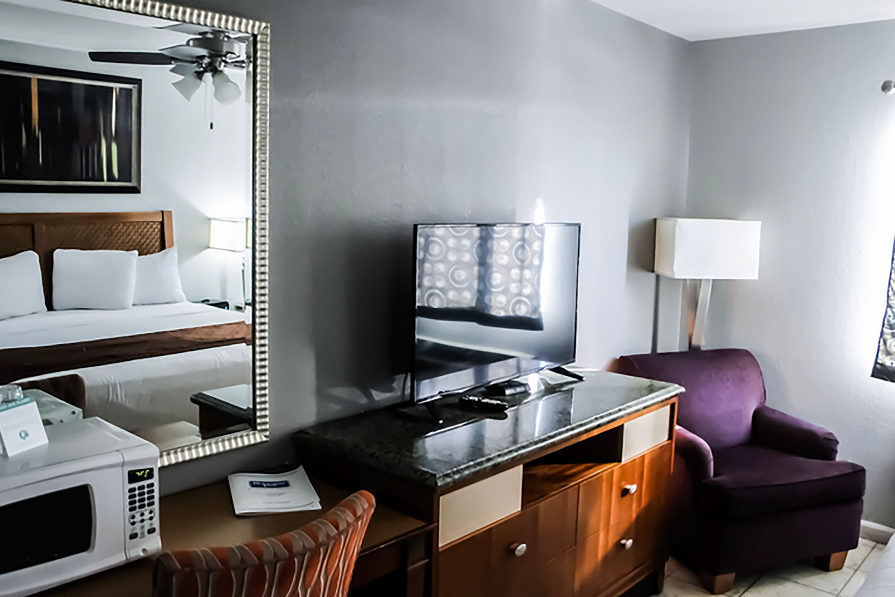 A hotel room with a bed, a desk with a television, a purple armchair, and a ceiling fan.