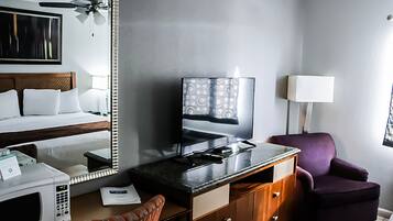 A hotel room with a bed, a desk with a television, a purple armchair, and a ceiling fan.