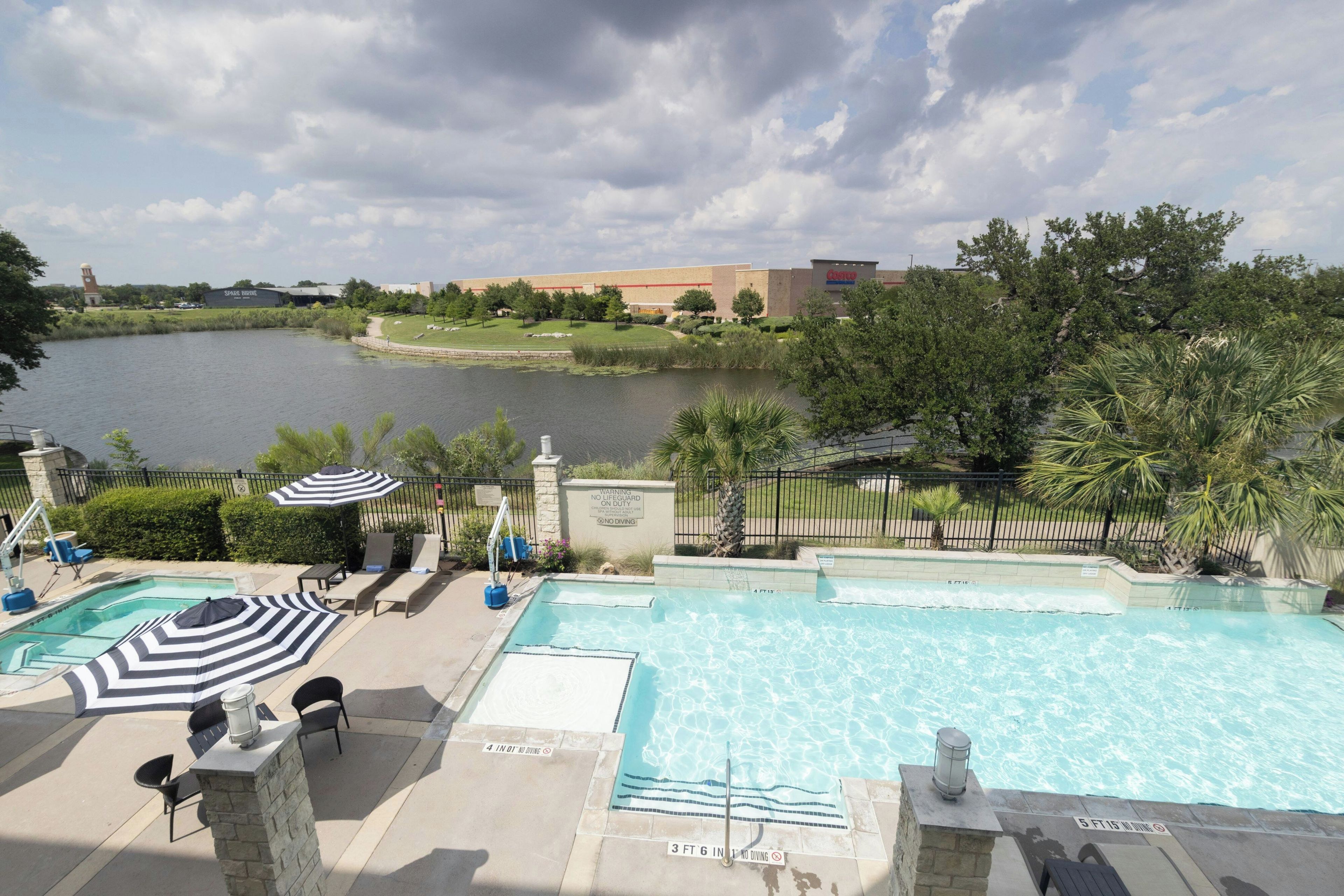 A swimming pool with lounge chairs and umbrellas, a warning sign, and a view of a lake and buildings in the background.