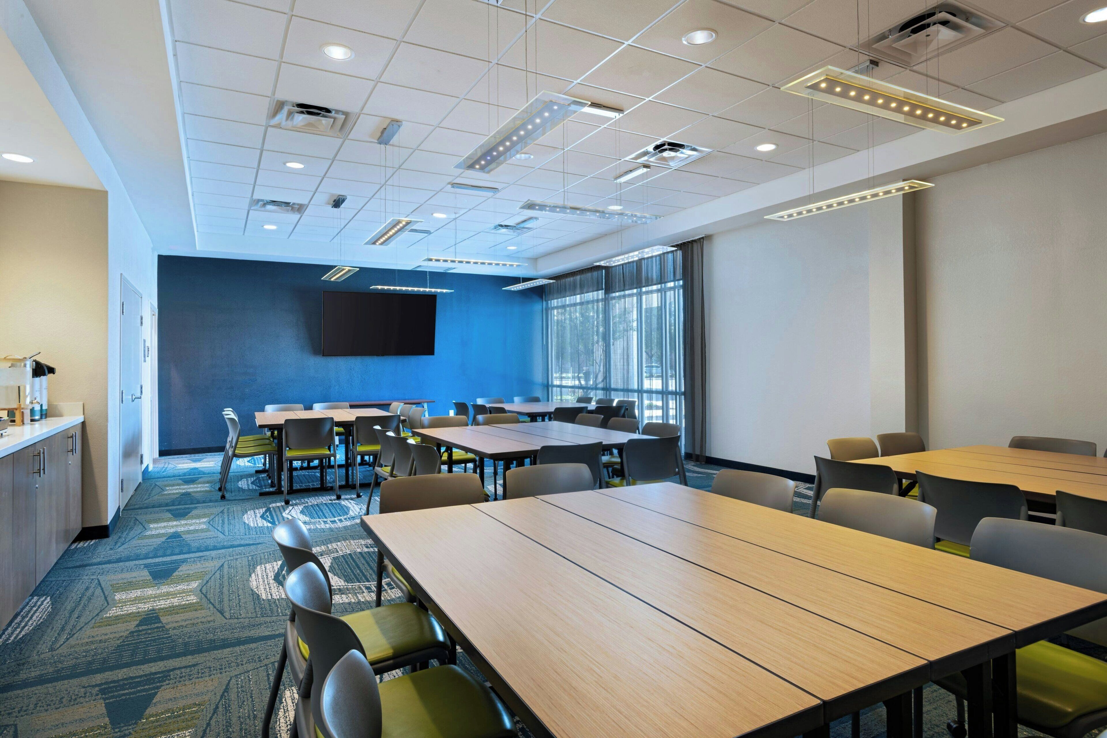 A conference room with multiple tables and chairs, a flat-screen TV mounted on the wall, and large windows providing natural light.