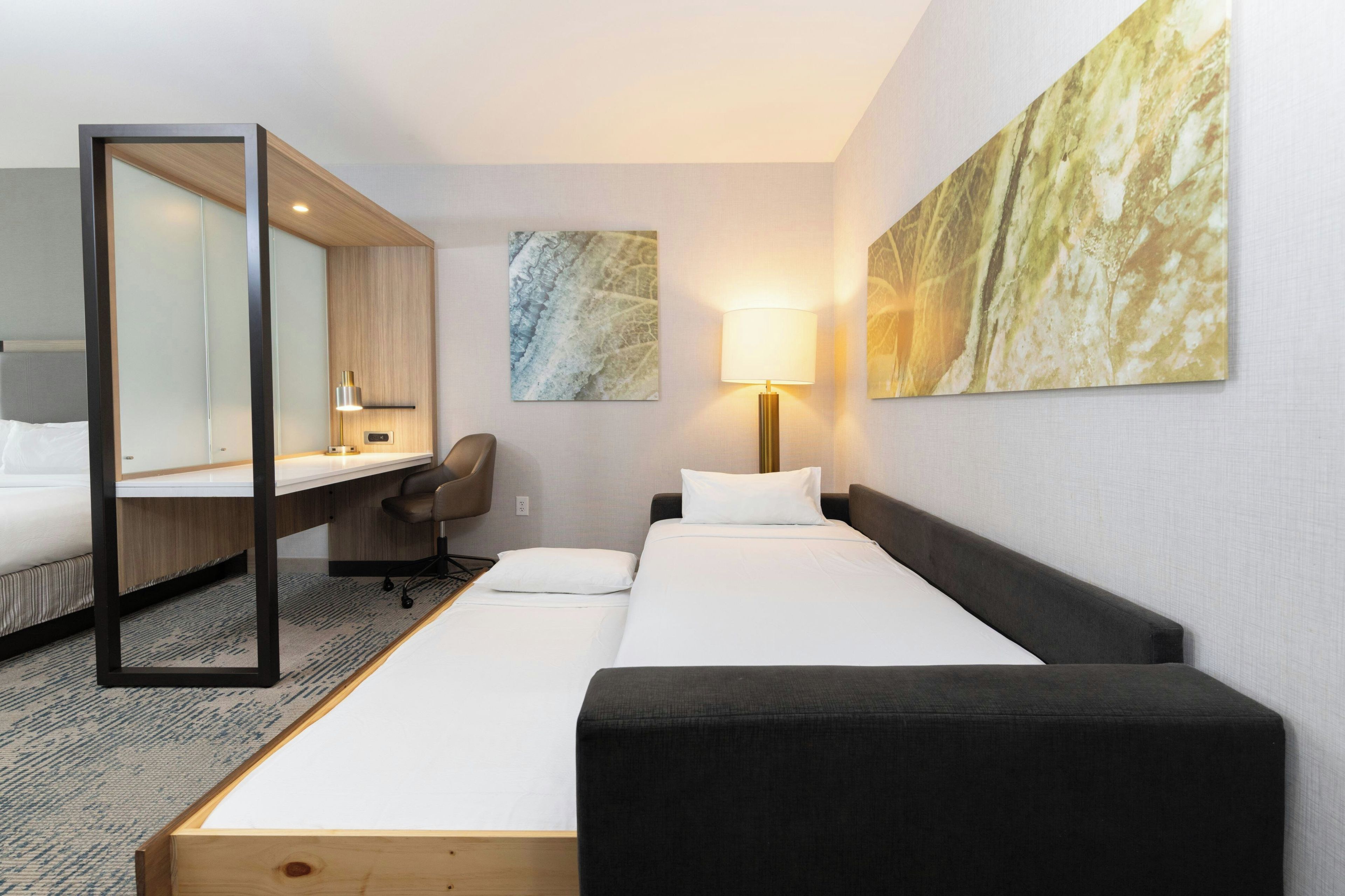 Habitación de hotel con cama, escritorio, silla, lámpara y una obra de arte en la pared.