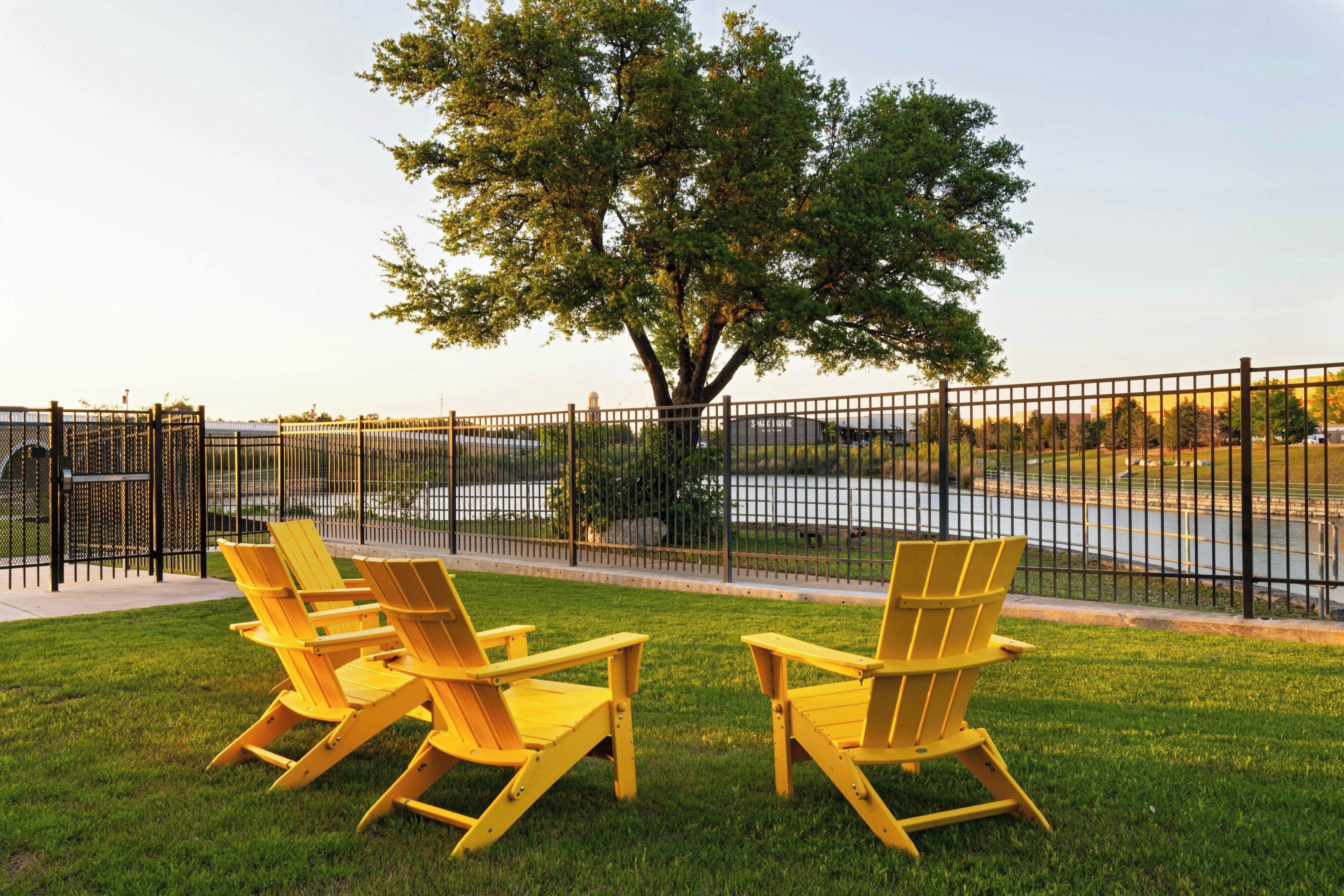 A fenced outdoor area with two yellow Adirondack chairs and a large tree.