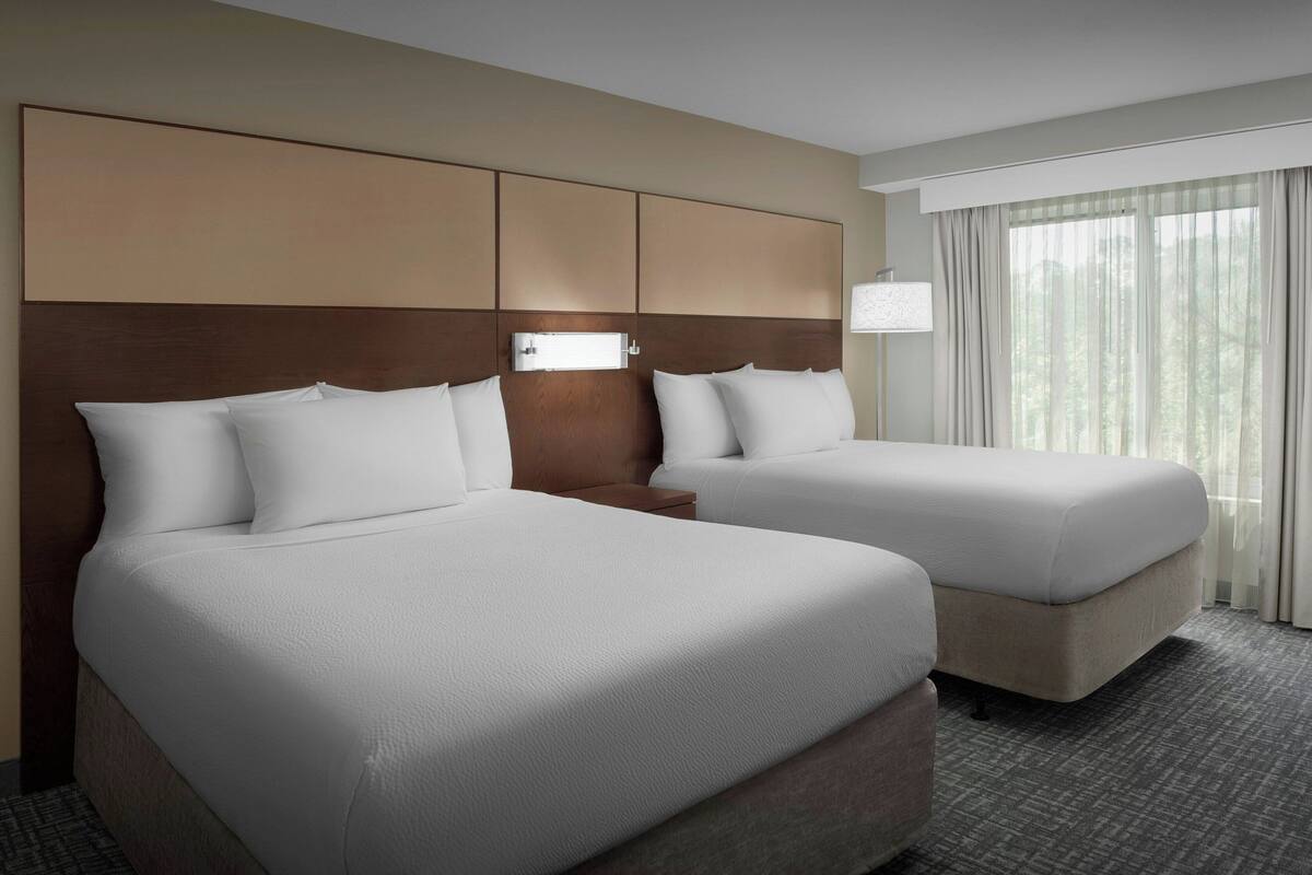 Une chambre d’hôtel avec deux lits, une tête de lit dotée de compartiments de rangement, une table de chevet et une fenêtre avec des rideaux.
