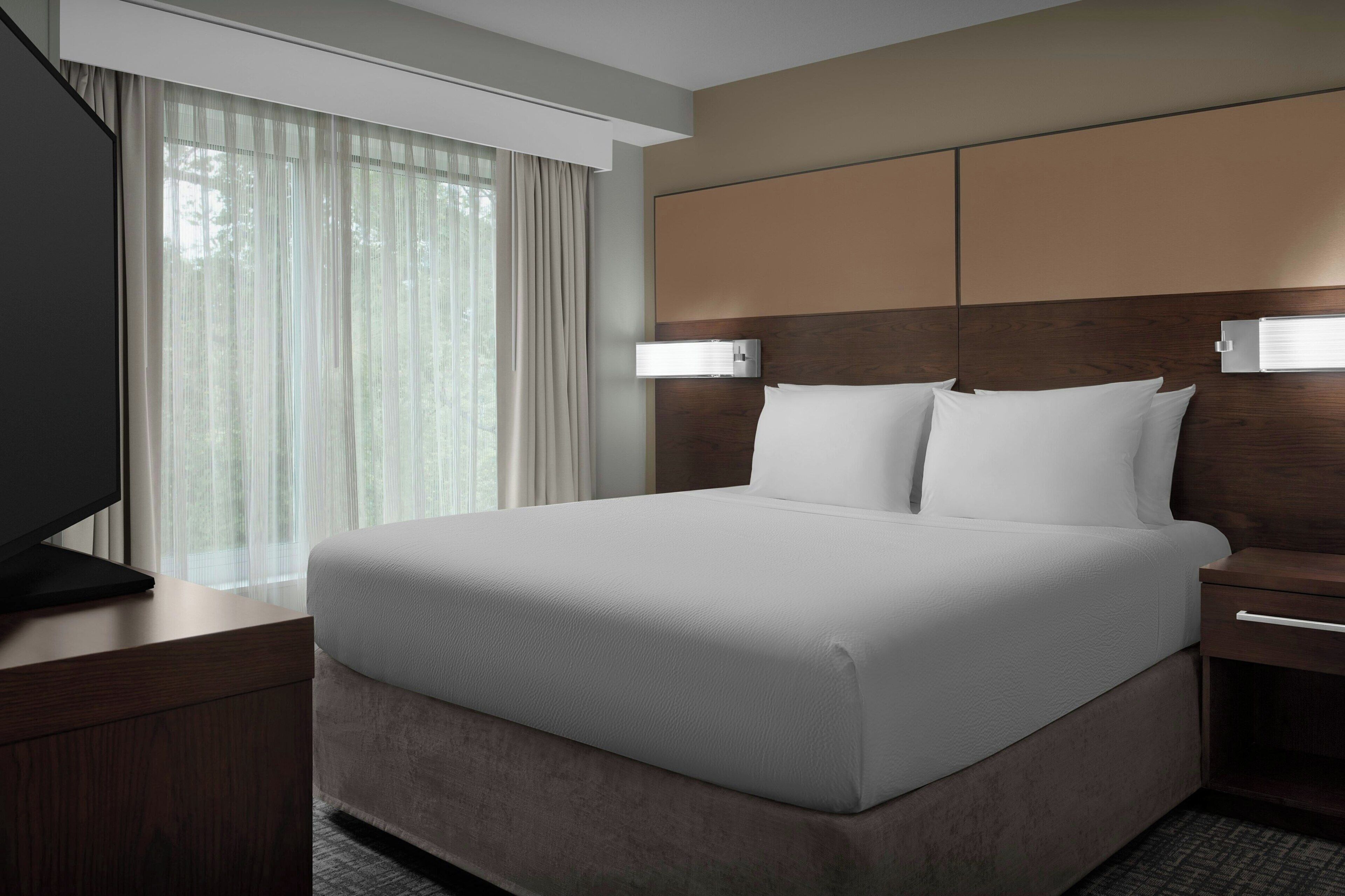 Une chambre d’hôtel avec un grand lit, un téléviseur posé sur une table de chevet en bois et une fenêtre ornée de rideaux légers.