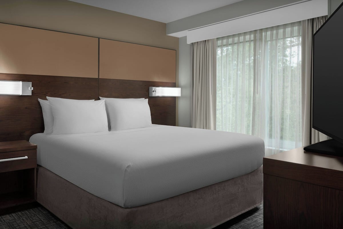Une chambre d’hôtel moderne dotée d’un grand lit, d’une tête de lit en bois, d’une table de chevet, d’un téléviseur à écran plat et d’une grande fenêtre avec des rideaux.