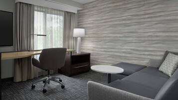 Una habitación de hotel moderna con un sofá gris, una pequeña mesa redonda, un escritorio con una silla, una lámpara y un amplio ventanal con cortinas.