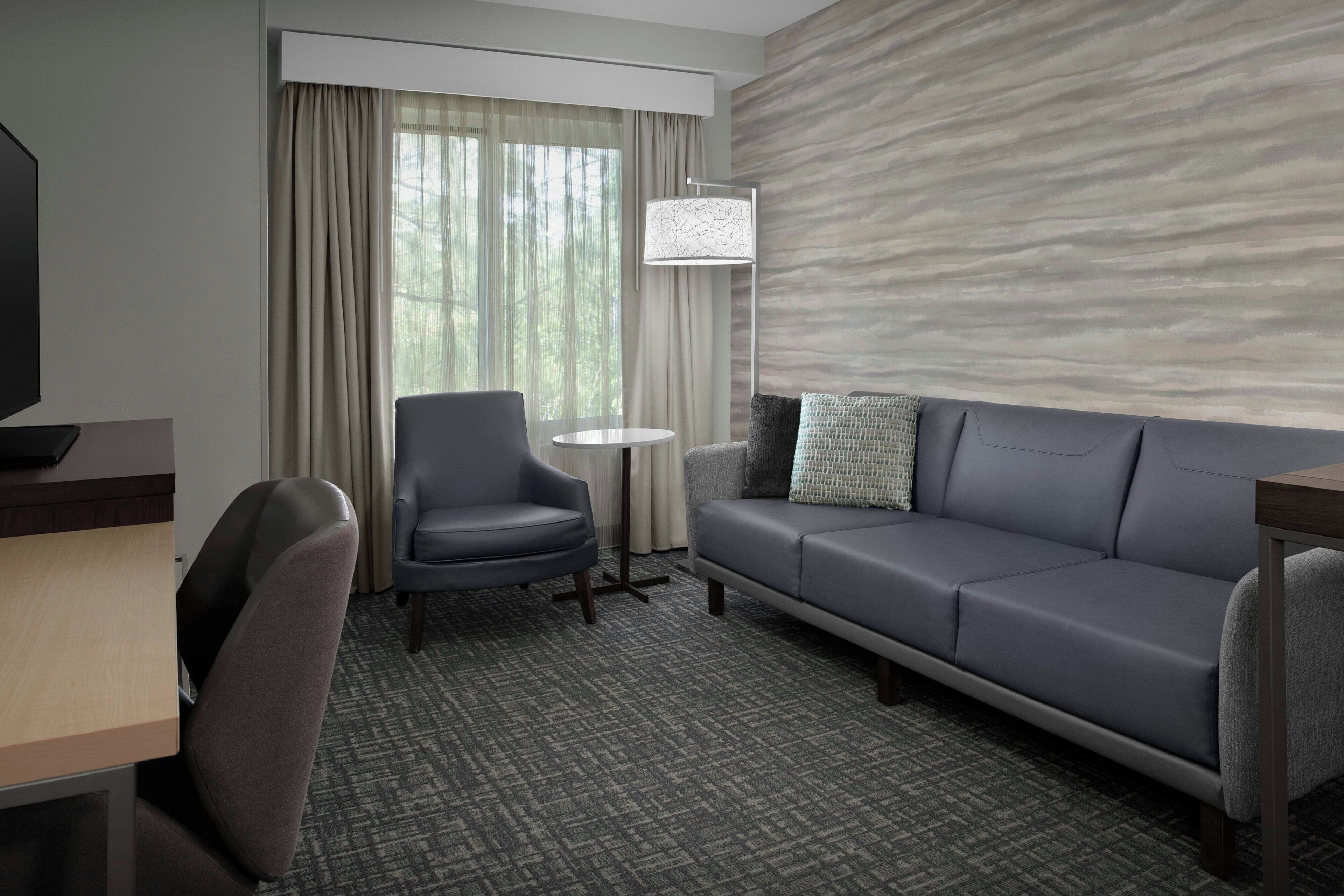 Habitación de hotel con sofá gris, una mesita redonda pequeña, un sillón gris, televisor y ventana con cortinas.