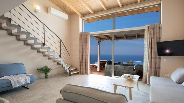Amplia sala de estar con escalera, un ventanal con vistas al mar, televisor de pantalla plana montado en la pared y un rincón de descanso acogedor.
