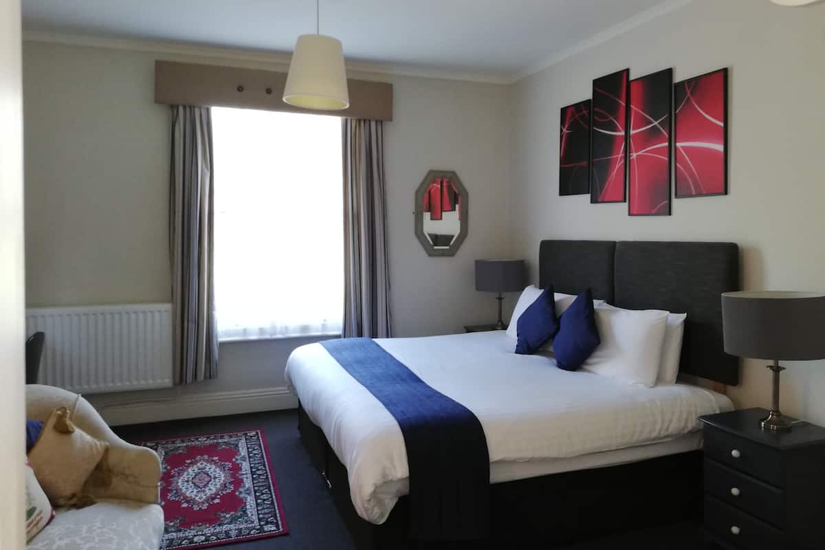 A hotel room with a large bed, a bedside table, a lamp, a window with curtains, and abstract wall art.