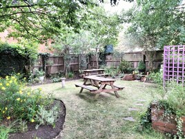 Un patio trasero con una mesa de picnic de madera rodeado de un jardín con diversas plantas y una cerca de madera.