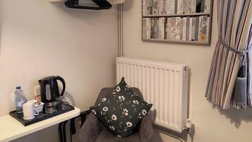 Een knus kamertje met een grijze fauteuil, een radiator, een boekenplank vol boeken, een plantje op een plank en een dienblad met een waterkoker, beker en waterfles.