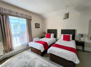 A hotel room with two beds, a large window with curtains, a hanging light fixture, and a small bedside table with a lamp. - The Claremont (Woodhall Spa)