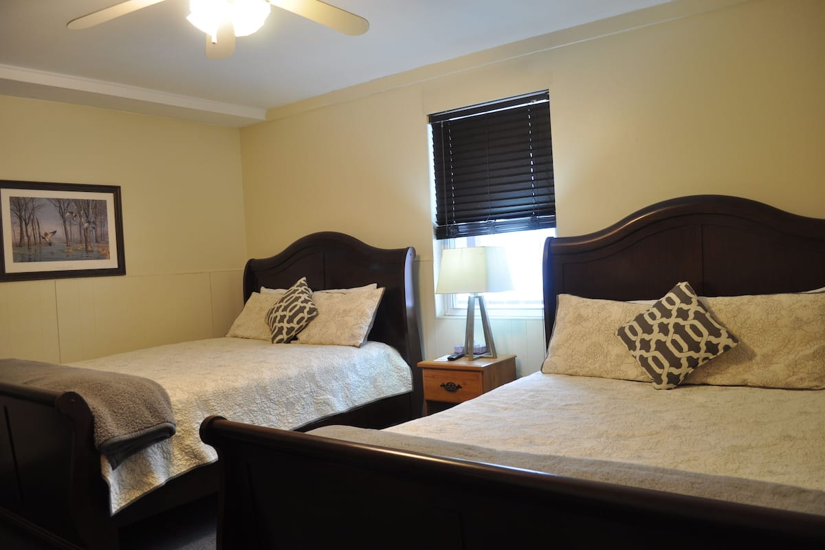 Une chambre avec deux lits simples, un ventilateur de plafond, un tableau encadré au mur et une fenêtre avec des stores.