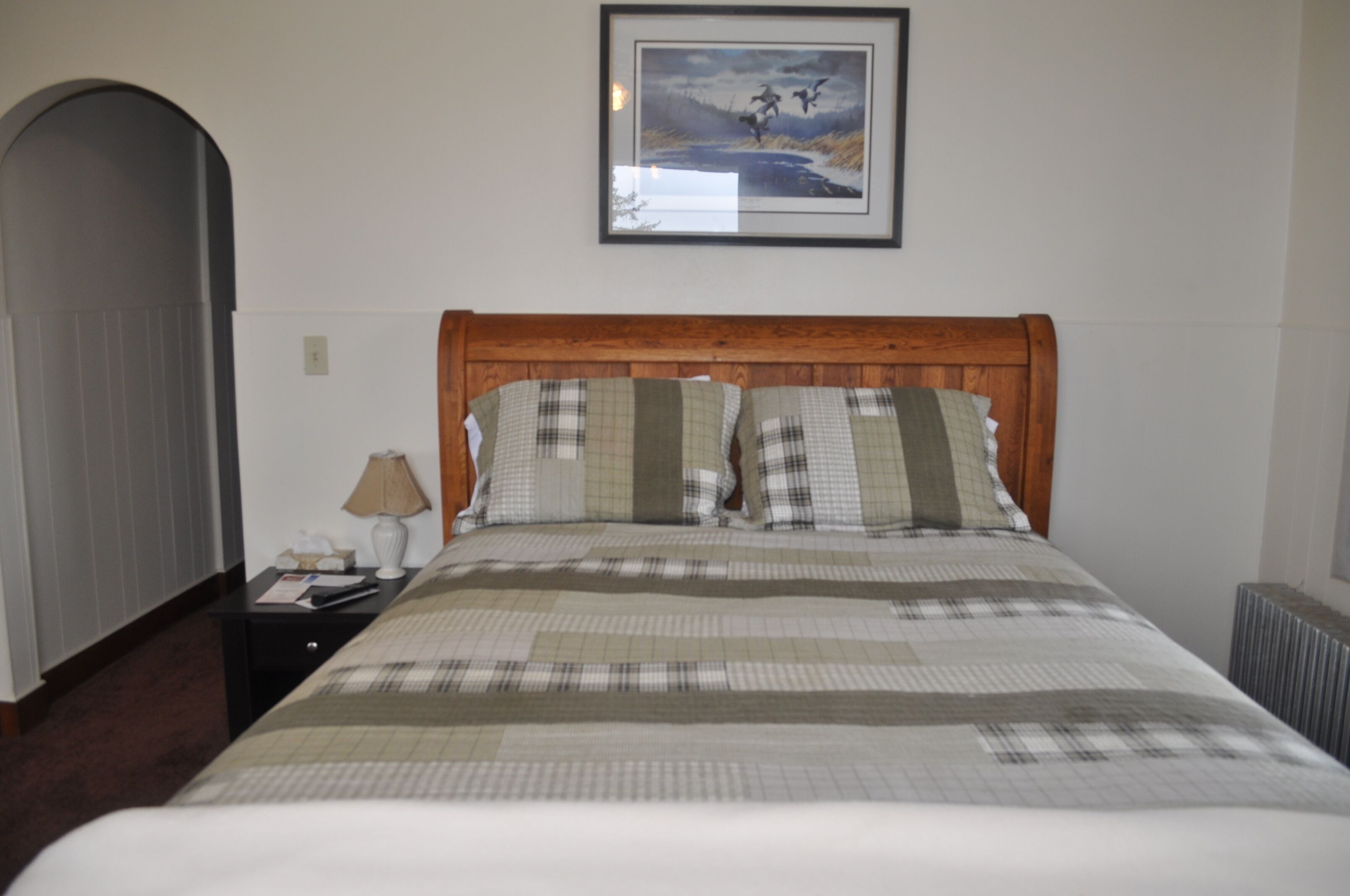 A neatly made bed with plaid bedding, a wooden headboard, a framed picture above the bed, a nightstand with a lamp, and a radiator on the floor.