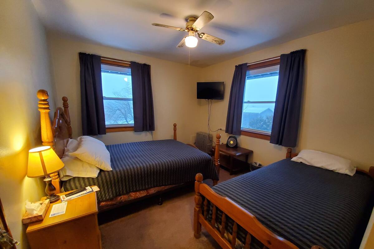 Une chambre d’hôtel avec deux lits simples, une table de chevet en bois avec une lampe, un ventilateur de plafond, un téléviseur fixé au mur et des fenêtres avec des rideaux.