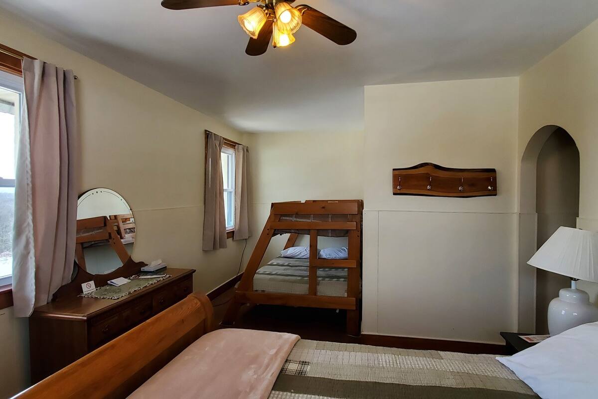Une chambre avec un lit en bois, un bureau, une chaise, un ventilateur de plafond, une fenêtre avec des rideaux et un porte-manteau fixé au mur.