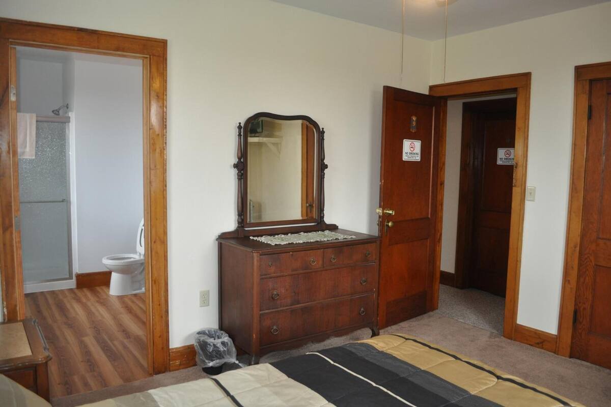 Une chambre à coucher avec une commode en bois, un miroir, un lit avec une literie à rayures et une salle de bain visible à travers une porte ouverte.