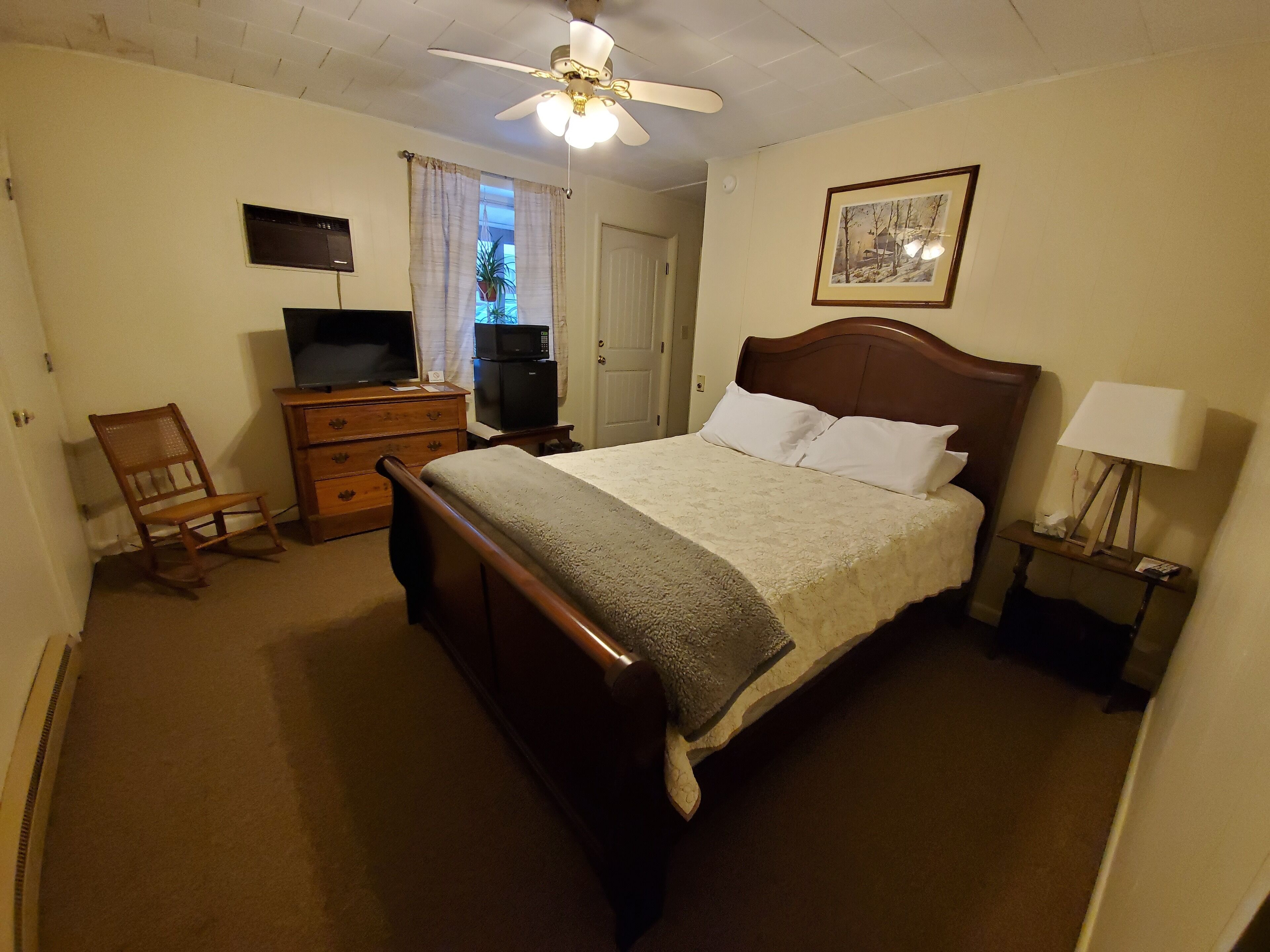 Quarto com cama de madeira, ventilador de teto, televisão, cômoda e abajur.