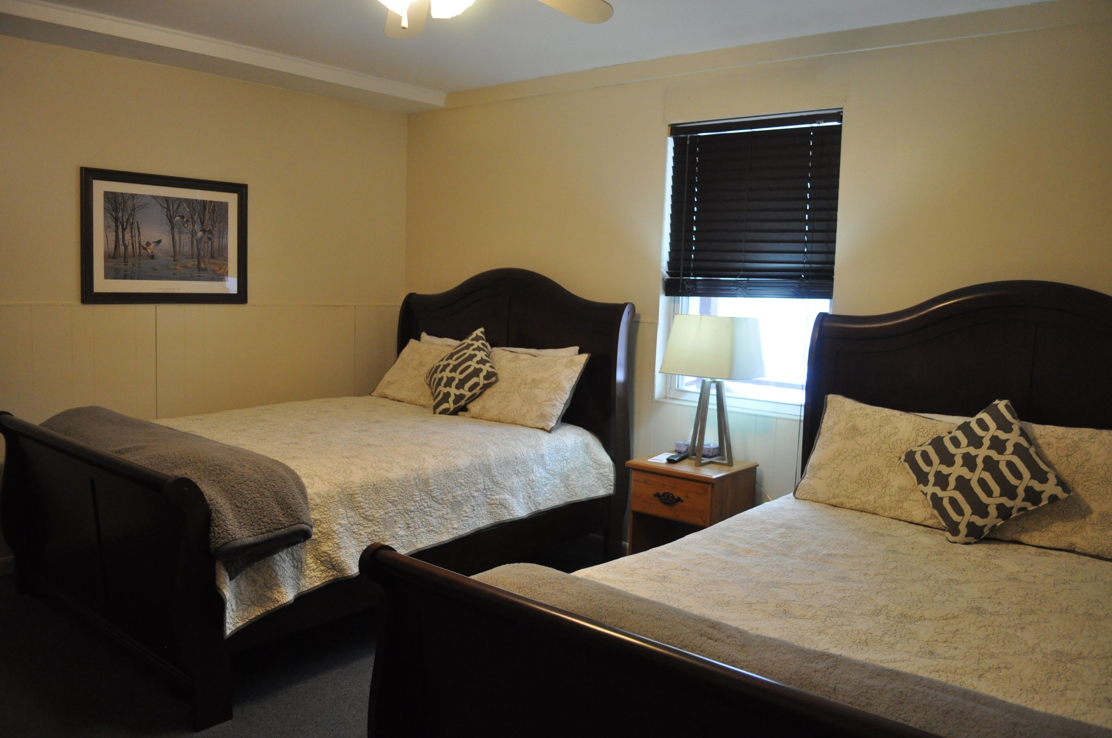 A bedroom with two single beds, a nightstand with a lamp, a framed picture on the wall, and a window with blinds.