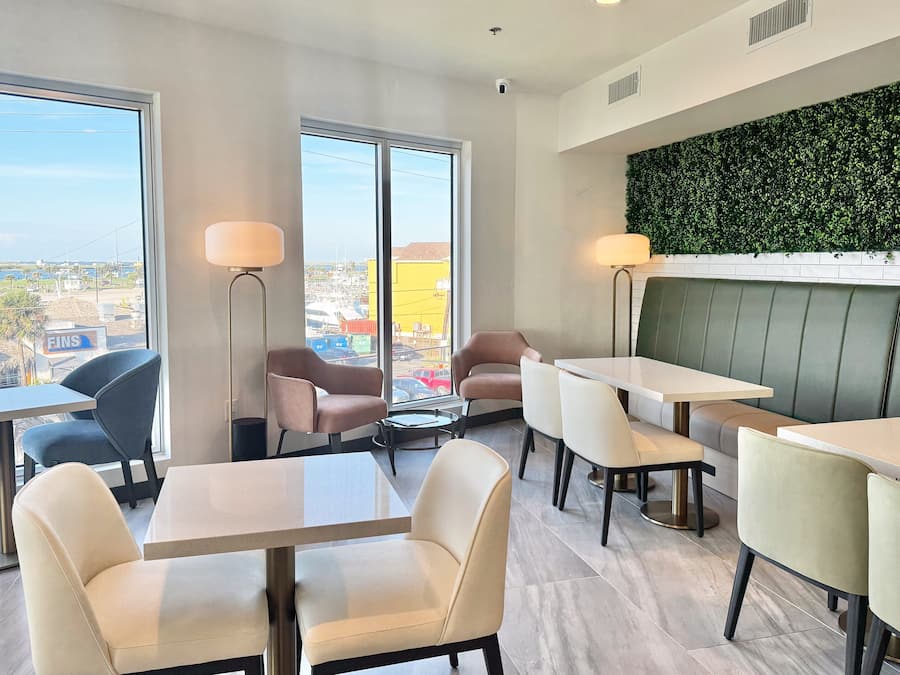 A modern hotel room with large windows, a green wall, and a seating area with tables and chairs.
