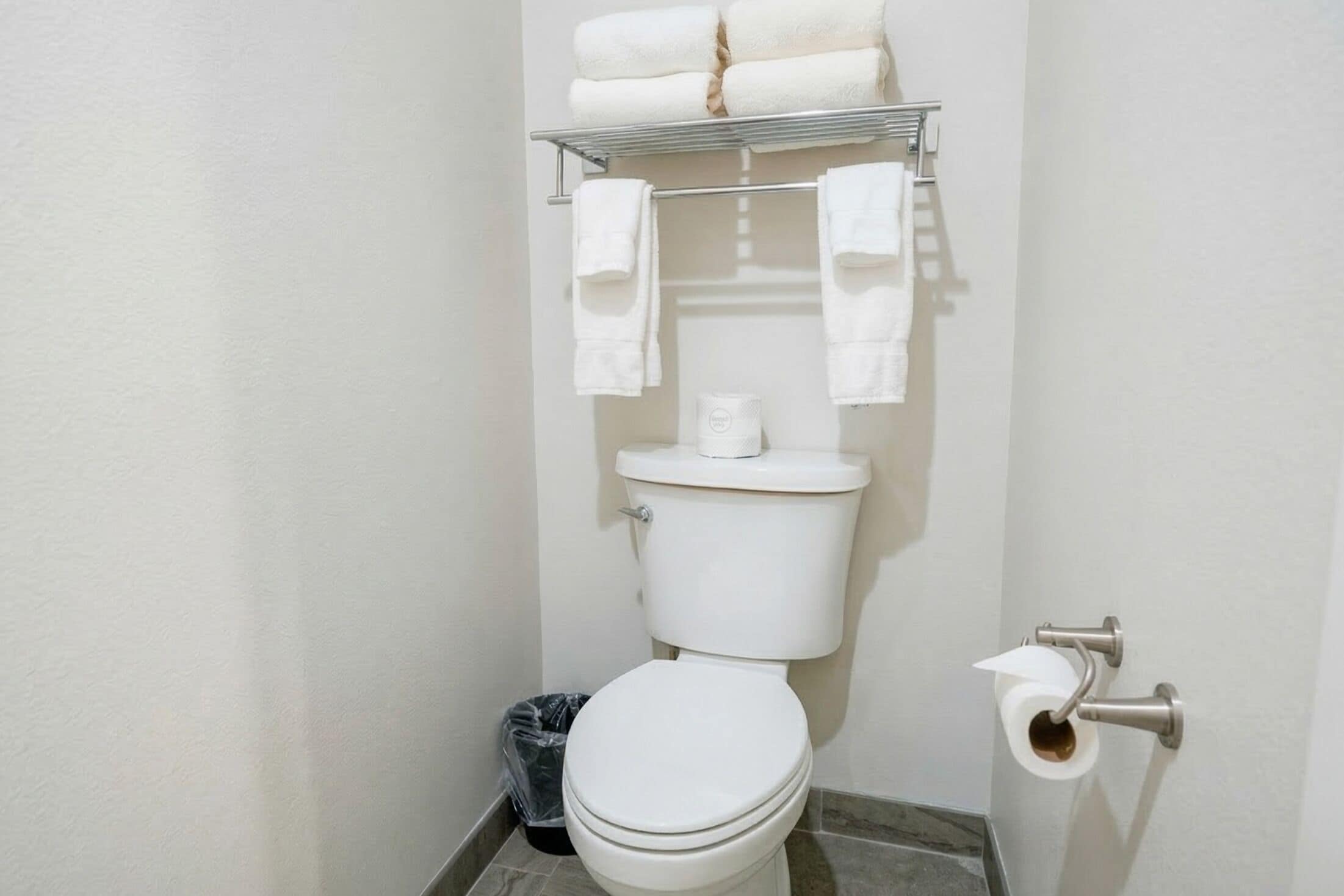 A bathroom with a toilet, a shelf with towels, a trash bin, and a toilet paper holder.
