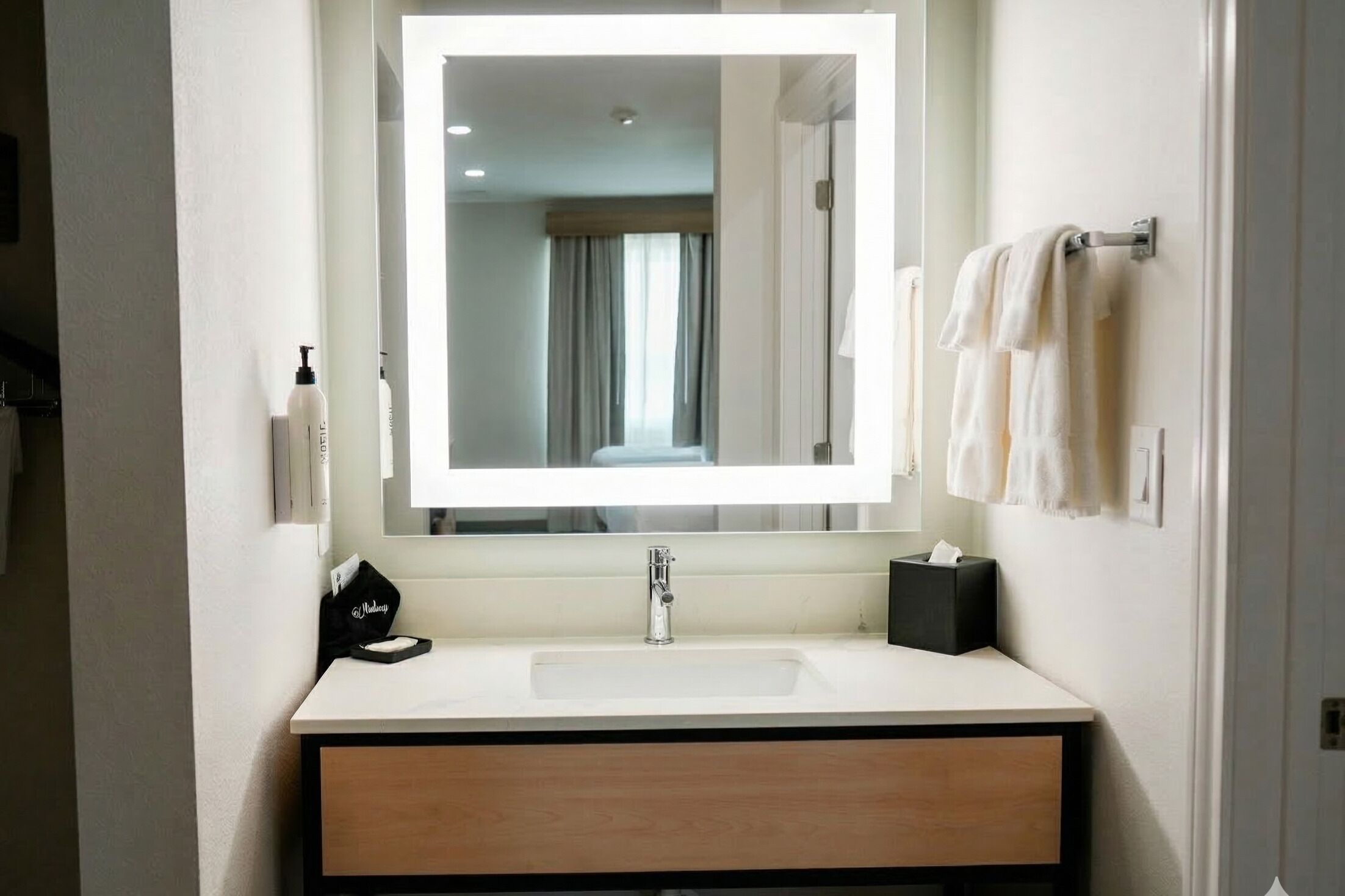 A hotel bathroom with a large mirror, a white sink, a black soap dispenser, and a towel rack with white towels.